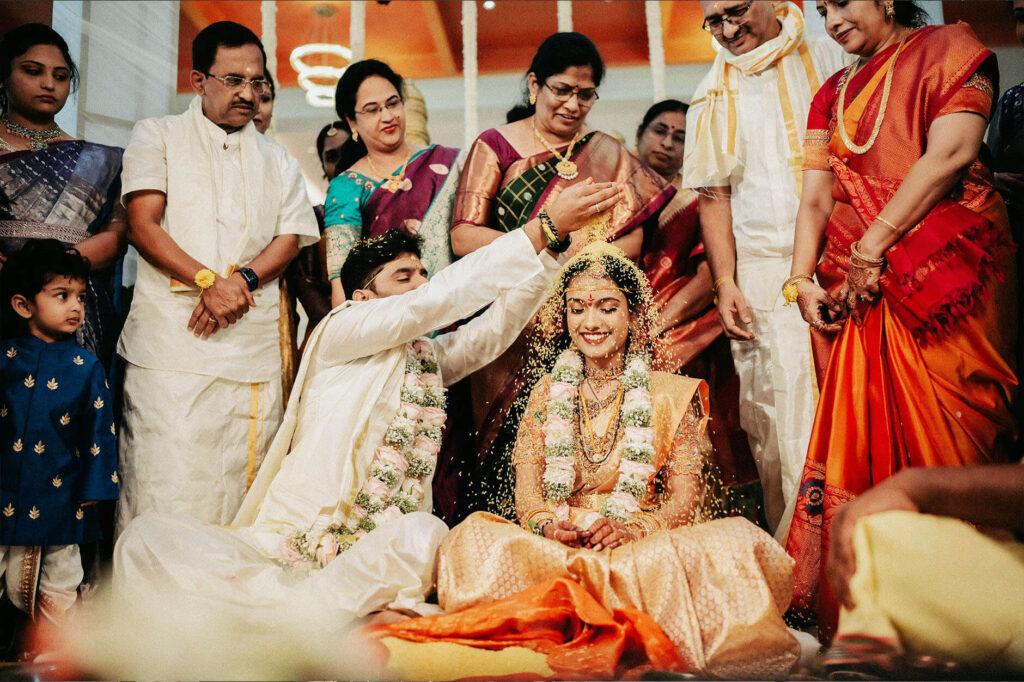 An Indian wedding ceremony with groom showering turmeric rice on the bride, surrounded by family.