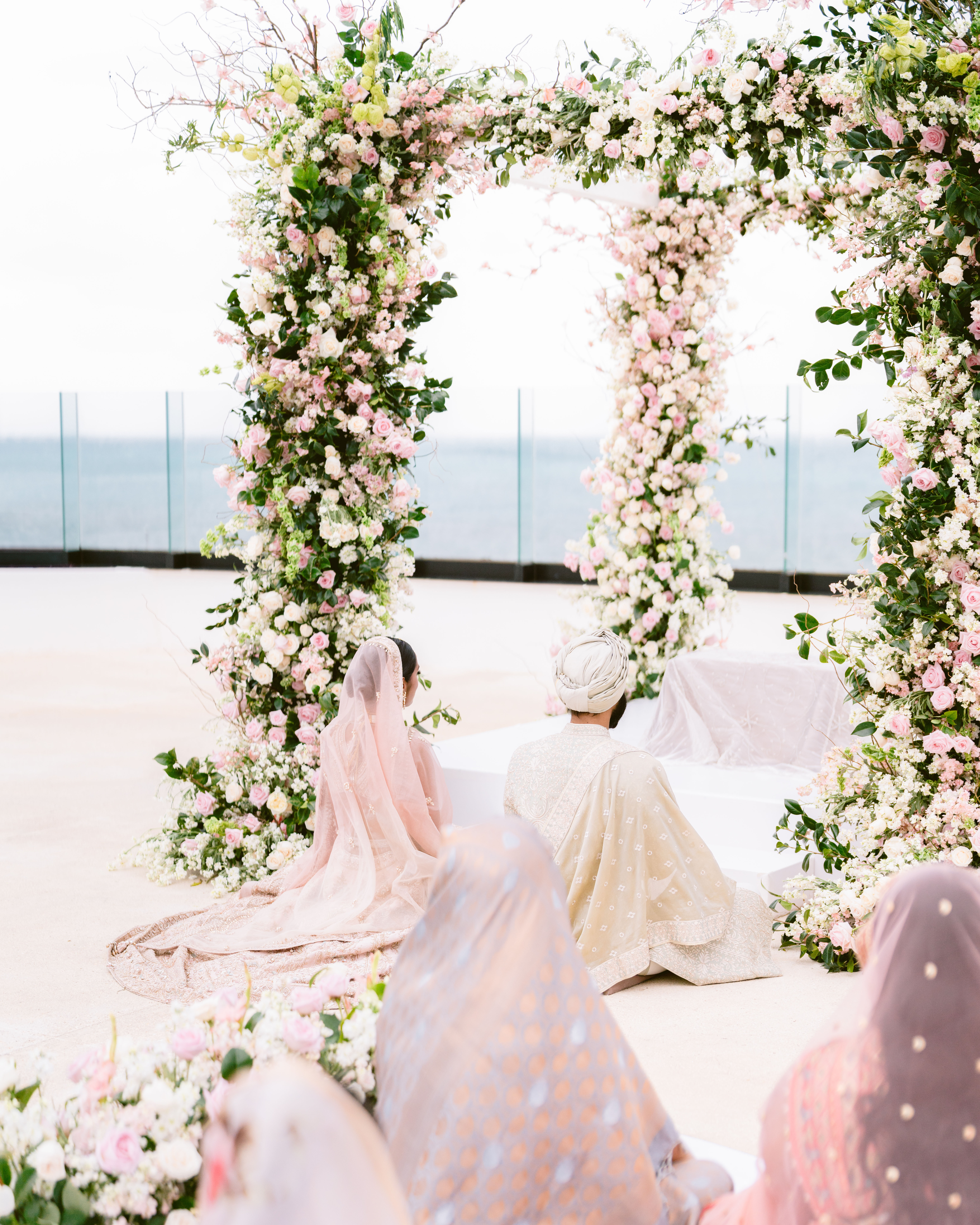 Sikh couple seated during an Anand Karaj ceremony under a floral canopy at an oceanfront destination wedding venue.