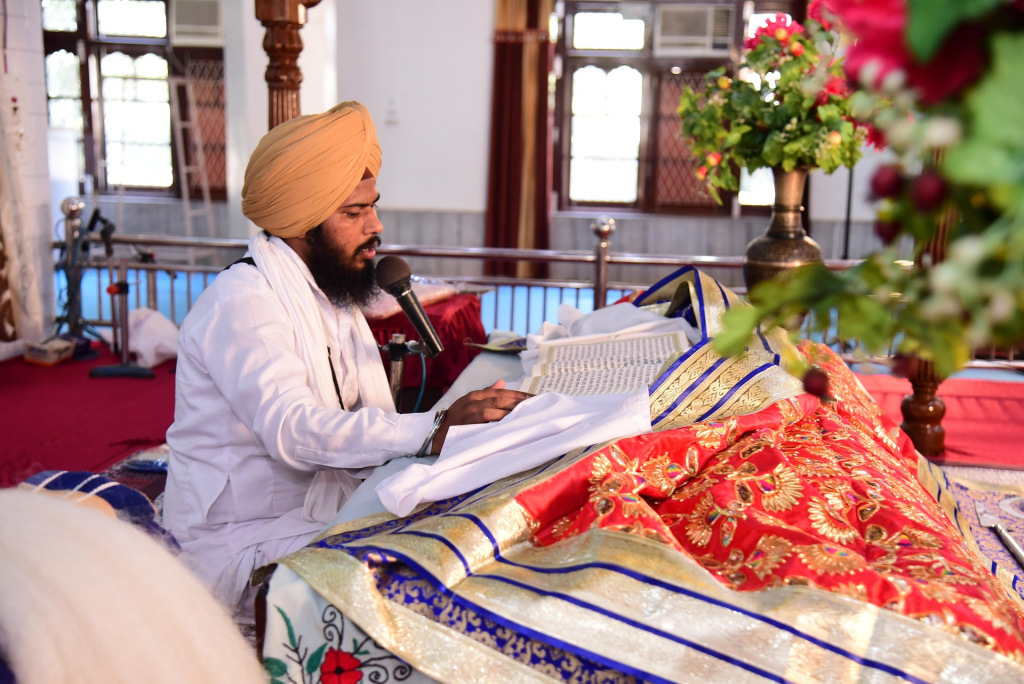 Granthi reciting from the Guru Granth Sahib during an Anand Karaj ceremony inside a Gurdwara, with traditional rumala and floral décor.